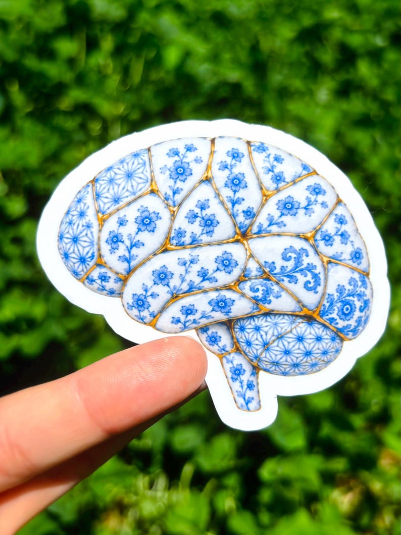 May include: A sticker shaped like a human brain featuring a blue and white floral pattern reminiscent of porcelain. The design includes gold-colored lines between the sections, mimicking the appearance of kintsugi repair on ceramic surfaces.