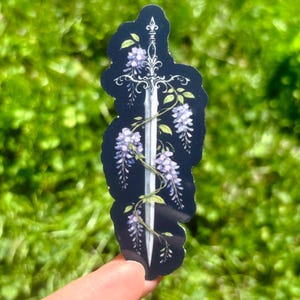 May include: A sticker featuring a silver sword design intertwined with purple wisteria flowers and green leaves against a dark blue background. The sticker has a black outline and is held up by a finger against a blurred green background.