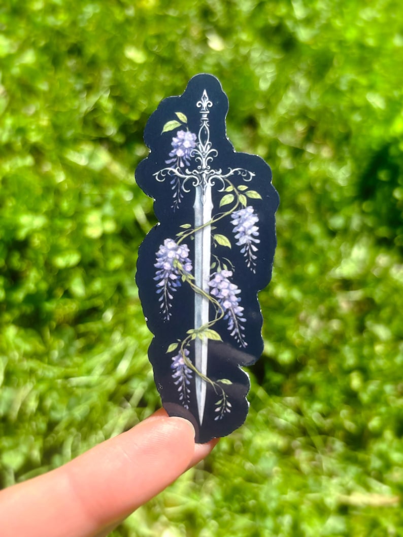May include: A sticker featuring a silver sword design intertwined with purple wisteria flowers and green leaves against a dark blue background. The sticker has a black outline and is held up by a finger against a blurred green background.