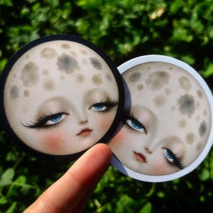 Peut inclure: Deux autocollants circulaires représentant un visage de lune stylisé au teint pâle, avec des taches de cratères gris foncé, des yeux bleu vif, de longs cils noirs et des joues roses. Un autocollant a une bordure noire, l'autre une bordure blanche.