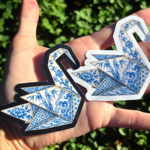 May include: Two origami crane stickers featuring a blue and white porcelain-inspired pattern. One sticker has a black border, while the other has a white border. The designs include intricate floral, bamboo, and wave motifs on the paper folds.