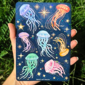 Pu&ograve; includere: Una carta blu scuro con illustrazioni di meduse colorate. Le meduse sono rosa, arancioni, verdi e blu, con accenti scintillanti e stelle. Il testo "PEACH CAT PRINTS" &egrave; visibile in basso.