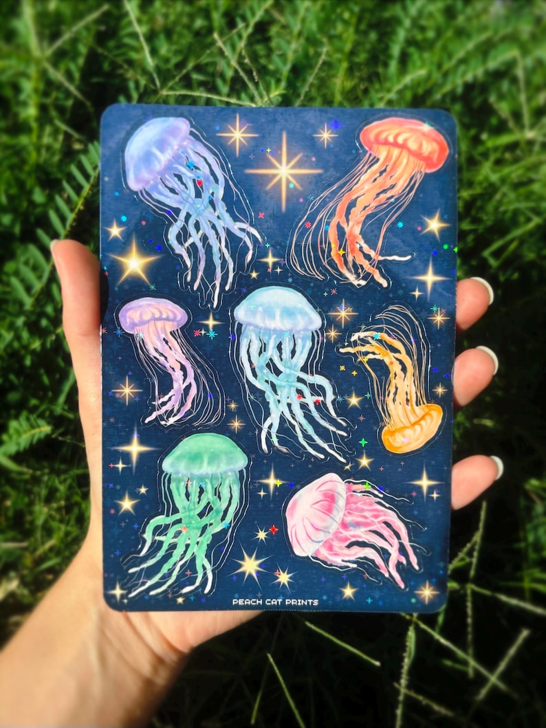 Pu&ograve; includere: Una carta blu scuro con illustrazioni di meduse colorate. Le meduse sono rosa, arancioni, verdi e blu, con accenti scintillanti e stelle. Il testo "PEACH CAT PRINTS" &egrave; visibile in basso.