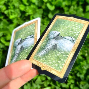 Puede incluir: Dos tarjetas rectangulares con marco dorado y borde negro. Las tarjetas presentan una ilustraci&oacute;n de una armadura tendida en un campo de flores blancas. El fondo es verde borroso.