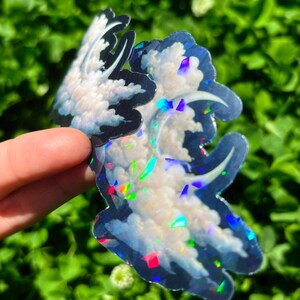May include: Two cloud-shaped stickers with a holographic, iridescent finish. The stickers feature a white cloud design with blue and black outlines. The stickers are held against a background of green foliage.