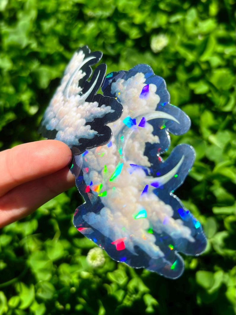May include: Two cloud-shaped stickers with a holographic, iridescent finish. The stickers feature a white cloud design with blue and black outlines. The stickers are held against a background of green foliage.