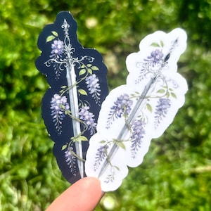 May include: Two stickers featuring a sword design intertwined with wisteria flowers. One sticker has a black background, the other is white. The sword is silver, and the flowers are purple and green. The stickers are held against a blurred green background.