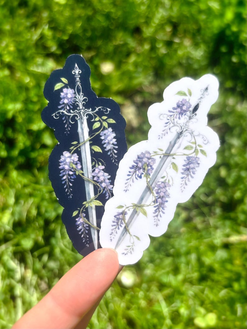 May include: Two stickers featuring a sword design intertwined with wisteria flowers. One sticker has a black background, the other is white. The sword is silver, and the flowers are purple and green. The stickers are held against a blurred green background.