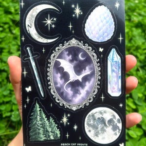 May include: A black sticker sheet featuring celestial and fantasy-themed illustrations. Designs include a crescent moon, stars, a dragon, a sword, a crystal, a full moon, and a dragon egg. The text "PEACH CAT PRINTS" is at the bottom.