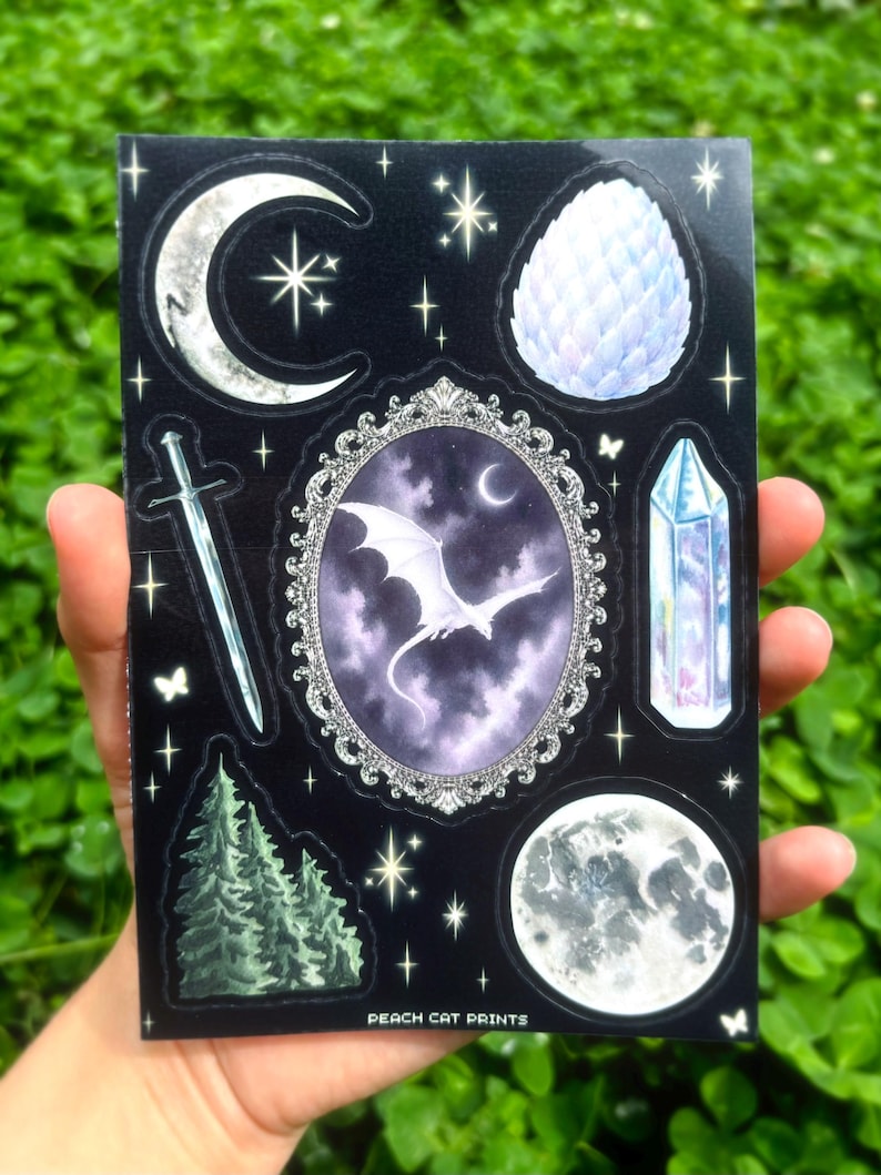 May include: A black sticker sheet featuring celestial and fantasy-themed illustrations. Designs include a crescent moon, stars, a dragon, a sword, a crystal, a full moon, and a dragon egg. The text "PEACH CAT PRINTS" is at the bottom.