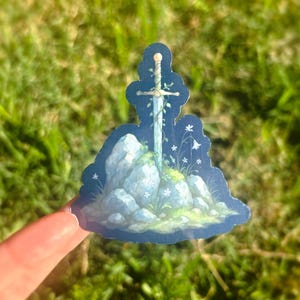 May include: A sticker featuring a sword embedded in a rock formation, with white flowers and green vines. The artwork has a fantasy theme, with a blue and white color palette. The sticker is held by a finger against a backdrop of green grass.