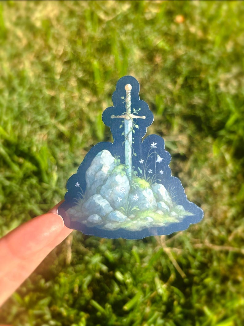 May include: A sticker featuring a sword embedded in a rock formation, with white flowers and green vines. The artwork has a fantasy theme, with a blue and white color palette. The sticker is held by a finger against a backdrop of green grass.