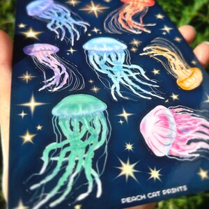 Pu&ograve; includere: Un foglio di adesivi blu scuro con illustrazioni di meduse colorate in tonalit&agrave; viola, blu, arancione, rosa e verde. Il design include stelle dorate e il testo "PEACH CAT PRINTS" in basso.