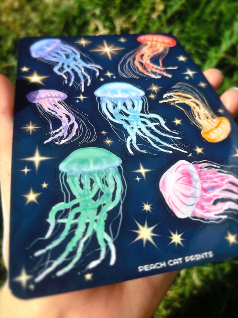 Pu&ograve; includere: Un foglio di adesivi blu scuro con illustrazioni di meduse colorate in tonalit&agrave; viola, blu, arancione, rosa e verde. Il design include stelle dorate e il testo "PEACH CAT PRINTS" in basso.