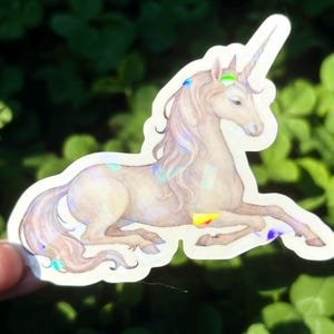 May include: A sticker featuring a light brown unicorn with a long, flowing mane and tail. The unicorn has a spiraled horn and is decorated with iridescent accents of blue, green, and yellow. The sticker is on a white background.