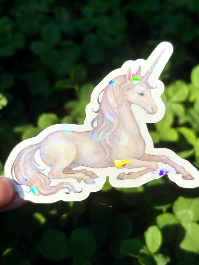 May include: A sticker featuring a light brown unicorn with a long, flowing mane and tail. The unicorn has a spiraled horn and is decorated with iridescent accents of blue, green, and yellow. The sticker is on a white background.