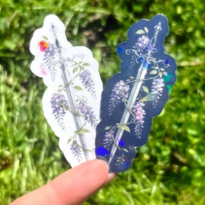 May include: Two stickers featuring a sword design with wisteria vines and flowers. One sticker is white, the other is dark blue. The stickers are held against a background of green grass.