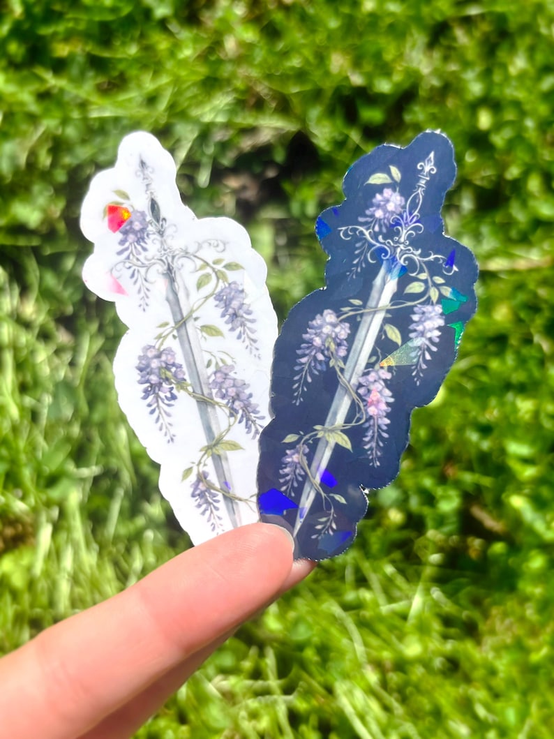 May include: Two stickers featuring a sword design with wisteria vines and flowers. One sticker is white, the other is dark blue. The stickers are held against a background of green grass.