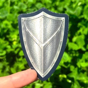 K&ouml;nnte beinhalten: Silberner Schildaufkleber mit schwarzem Rand. Der Schild hat eine strukturierte Oberfl&auml;che und ein stilisiertes Design. Der Aufkleber wird vor einem Hintergrund aus gr&uuml;nem Laub gehalten.