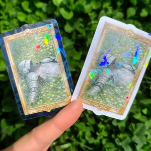 Puede incluir: Dos pegatinas con una armadura de caballero en un campo de flores, enmarcadas en oro. Las pegatinas tienen un efecto hologr&aacute;fico, con colores del arco&iacute;ris visibles. El fondo es un campo de follaje verde.