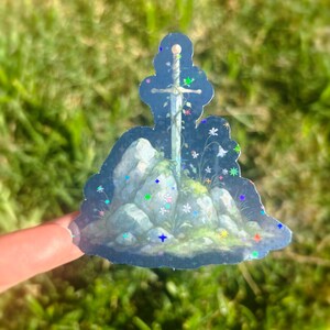 May include: A holographic sticker featuring a sword embedded in a rock formation with colorful floral accents. The sticker has a blue background with glittery stars and is held against a blurred green background.