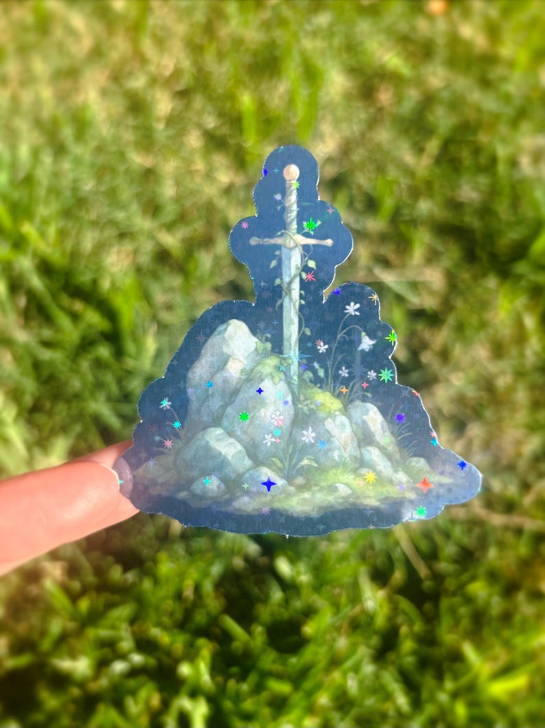 May include: A holographic sticker featuring a sword embedded in a rock formation with colorful floral accents. The sticker has a blue background with glittery stars and is held against a blurred green background.