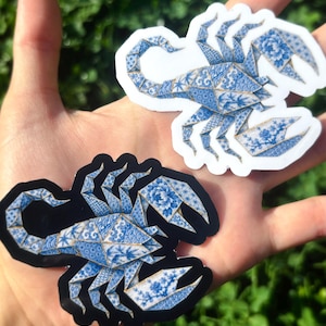 May include: Two scorpion-shaped stickers featuring a blue and white porcelain-inspired pattern with intricate floral and geometric motifs. One sticker has a white border, while the other has a black border, both showcasing a segmented, angular design.