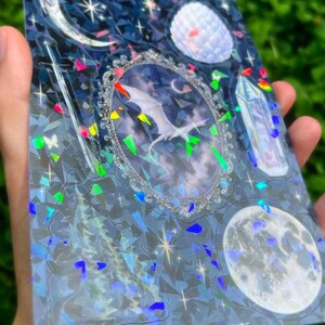 May include: A holographic sticker sheet featuring celestial and fantasy elements. The design includes a central framed image of a dragon, moons, stars, and a crystal. The text "PEACH CAT PRINTS" is visible at the bottom.