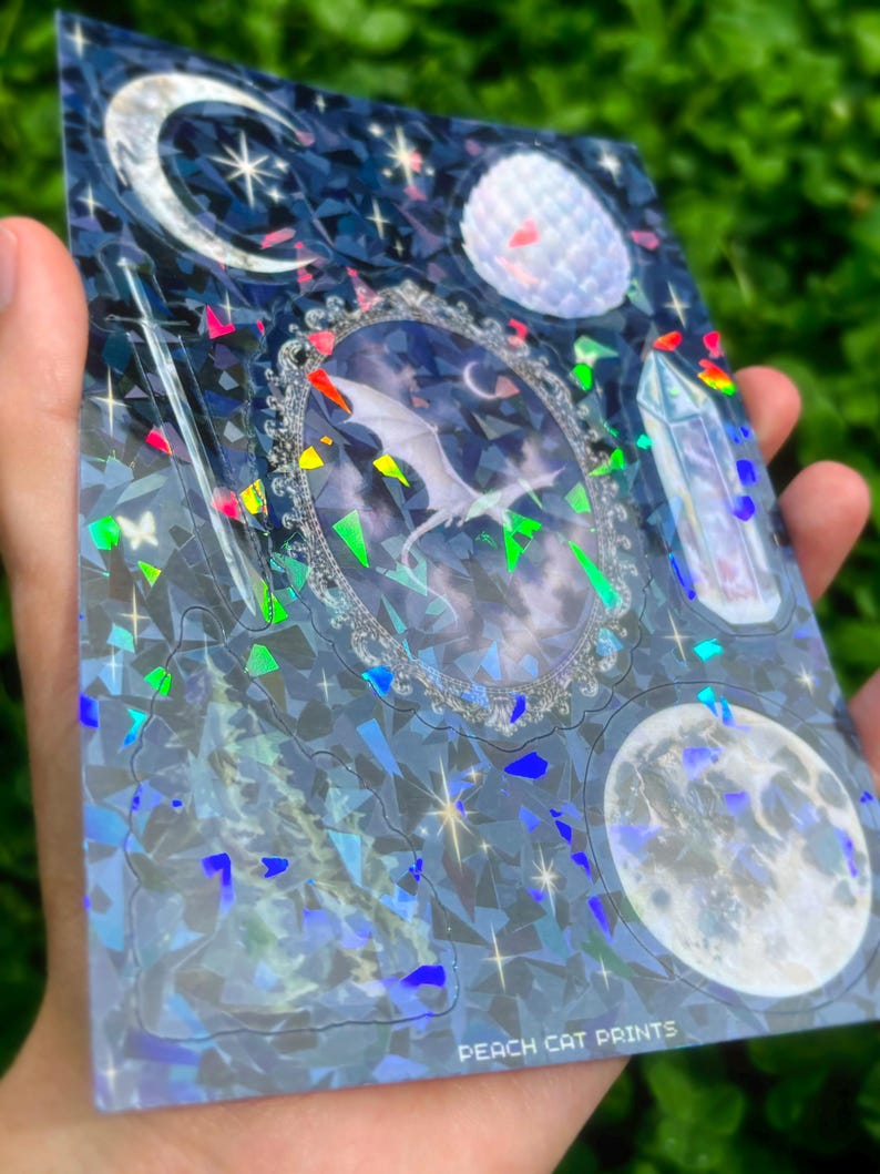 May include: A holographic sticker sheet featuring celestial and fantasy elements. The design includes a central framed image of a dragon, moons, stars, and a crystal. The text "PEACH CAT PRINTS" is visible at the bottom.