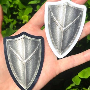 K&ouml;nnte beinhalten: Zwei schildf&ouml;rmige Aufkleber mit metallisch-silbernem Finish. Einer befindet sich auf schwarzem Hintergrund, der andere hat einen wei&szlig;en Rand. Beide haben eine strukturierte Oberfl&auml;che und ein zentrales Designelement.