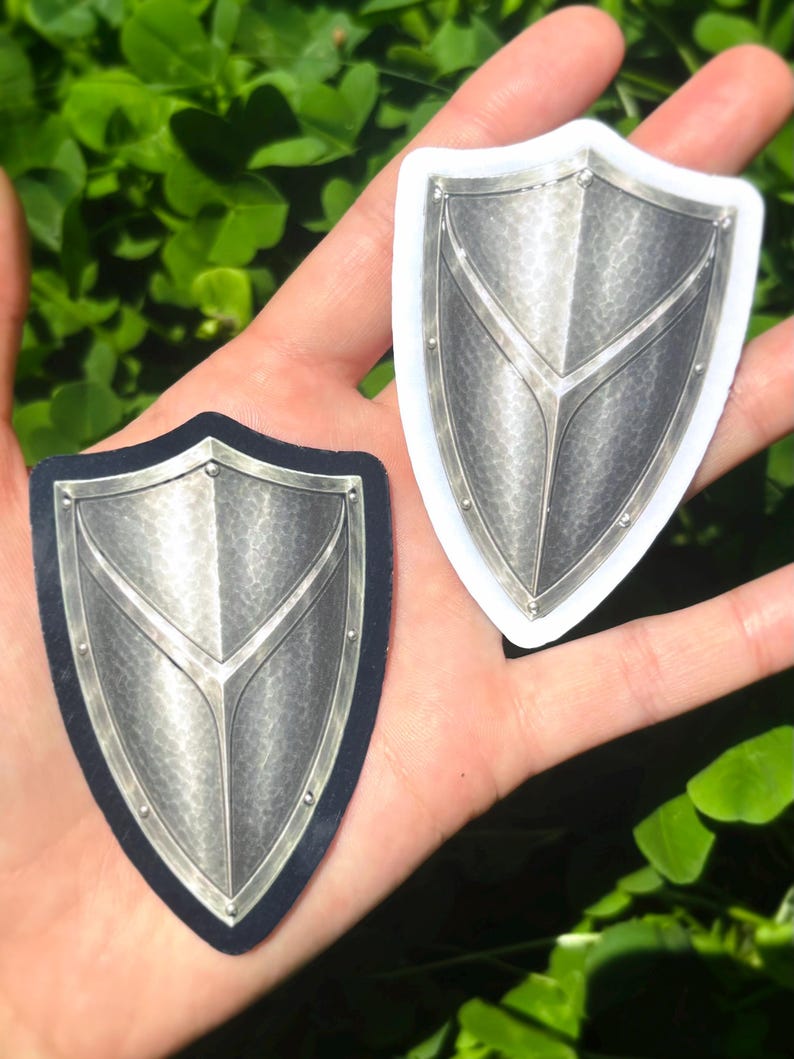 K&ouml;nnte beinhalten: Zwei schildf&ouml;rmige Aufkleber mit metallisch-silbernem Finish. Einer befindet sich auf schwarzem Hintergrund, der andere hat einen wei&szlig;en Rand. Beide haben eine strukturierte Oberfl&auml;che und ein zentrales Designelement.