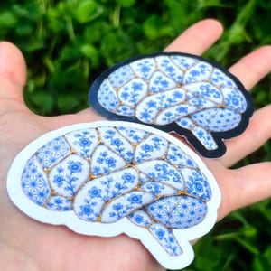 May include: Two brain-shaped stickers featuring a white and blue floral pattern reminiscent of porcelain, with gold-colored lines separating the sections. One sticker has a white border, while the other has a black border, both showing anatomical detail.