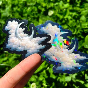 May include: Two cloud-shaped stickers with a crescent moon design. The clouds are white with blue and black outlines. One sticker has a holographic effect with rainbow colors. The stickers are held against a green background.