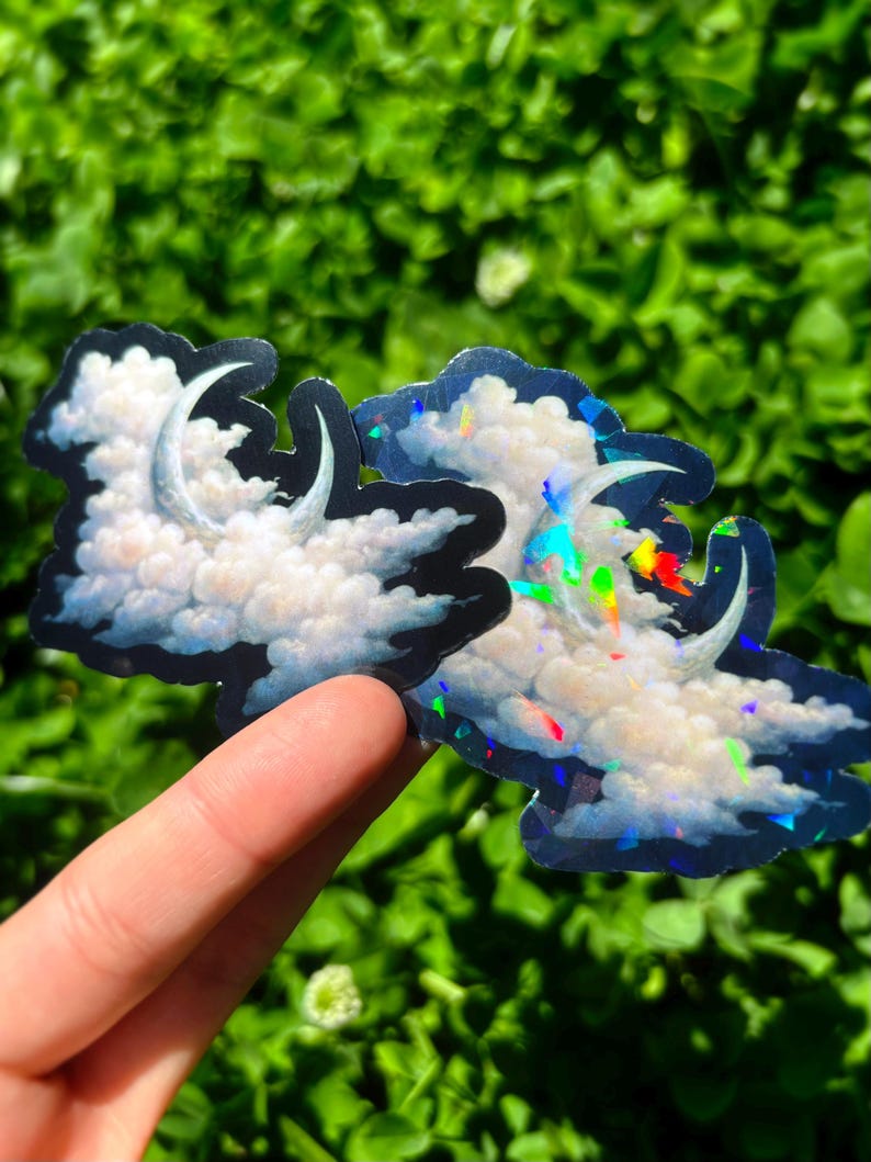 May include: Two cloud-shaped stickers with a crescent moon design. The clouds are white with blue and black outlines. One sticker has a holographic effect with rainbow colors. The stickers are held against a green background.