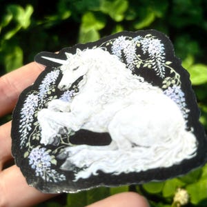 Puede incluir: Una pegatina de unicornio blanco con borde negro, rodeada de flores moradas claras y hojas verdes. El unicornio est&aacute; tumbado con la cabeza levantada y su cuerno es visible. La pegatina se sostiene en una mano sobre un fondo de follaje verde.