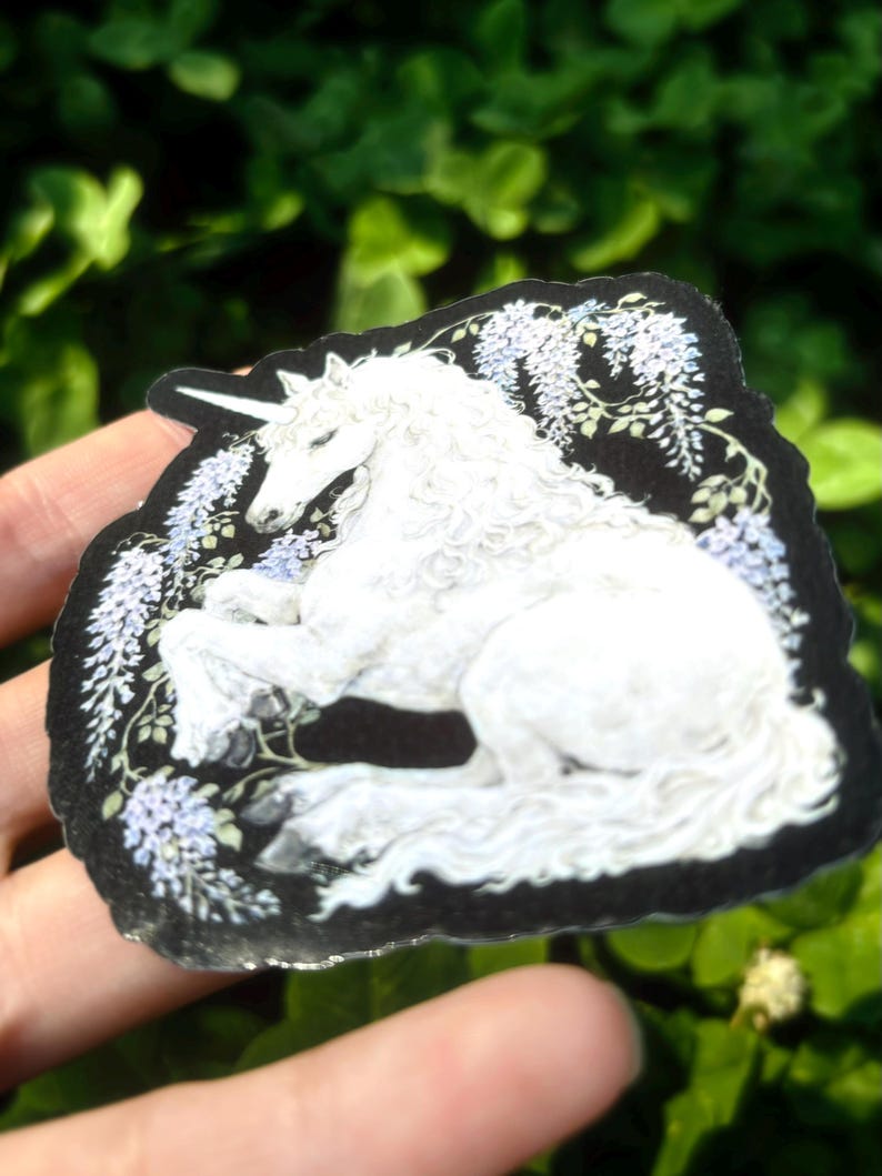 Puede incluir: Una pegatina de unicornio blanco con borde negro, rodeada de flores moradas claras y hojas verdes. El unicornio est&aacute; tumbado con la cabeza levantada y su cuerno es visible. La pegatina se sostiene en una mano sobre un fondo de follaje verde.