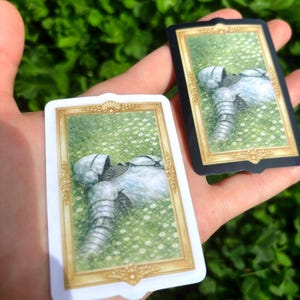 Puede incluir: Dos cartas que presentan una ilustraci&oacute;n de una armadura medieval descansando en un campo de flores blancas. Una carta tiene un borde blanco con detalles dorados, la otra tiene un borde negro con detalles dorados. El fondo es verde borroso.