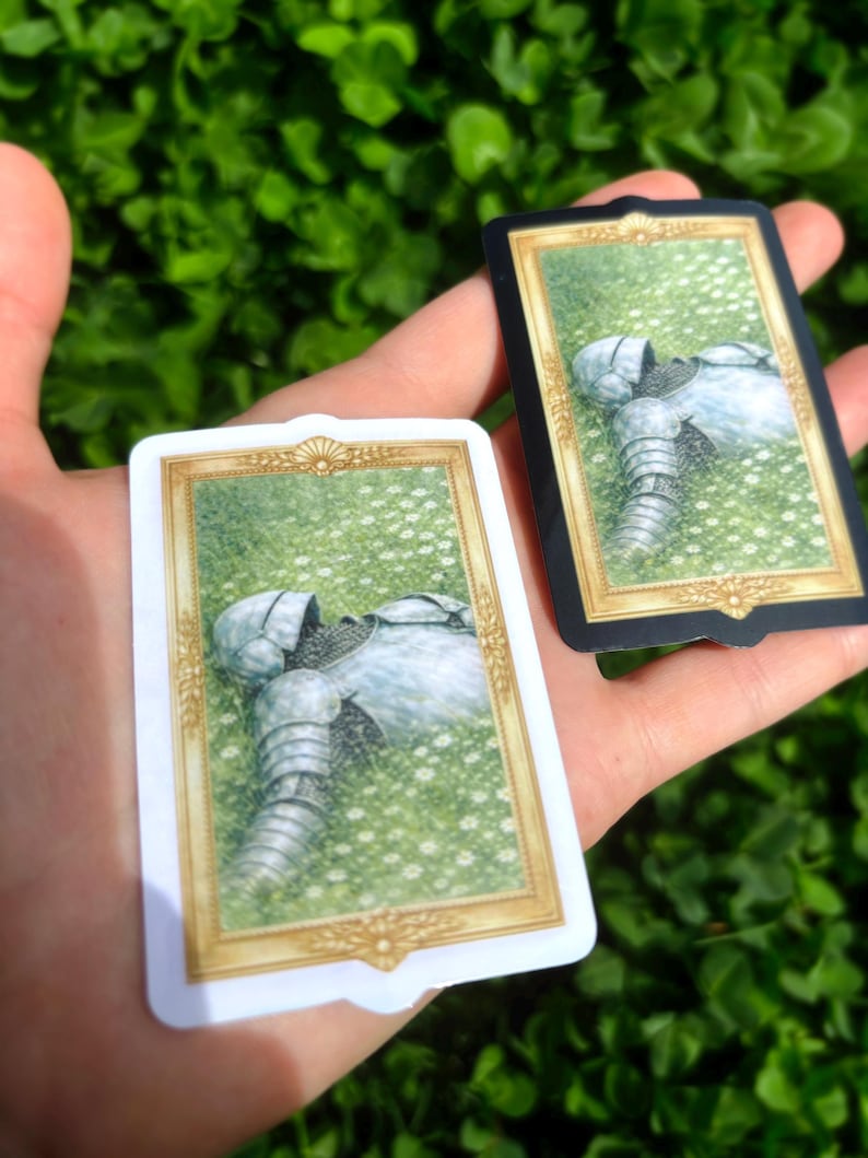 Puede incluir: Dos cartas que presentan una ilustraci&oacute;n de una armadura medieval descansando en un campo de flores blancas. Una carta tiene un borde blanco con detalles dorados, la otra tiene un borde negro con detalles dorados. El fondo es verde borroso.