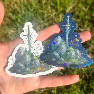 May include: Two stickers featuring a sword in a stone design. The stickers depict a sword embedded in a rock formation with floral accents. One sticker has a white background, and the other has a dark blue background with holographic glitter.