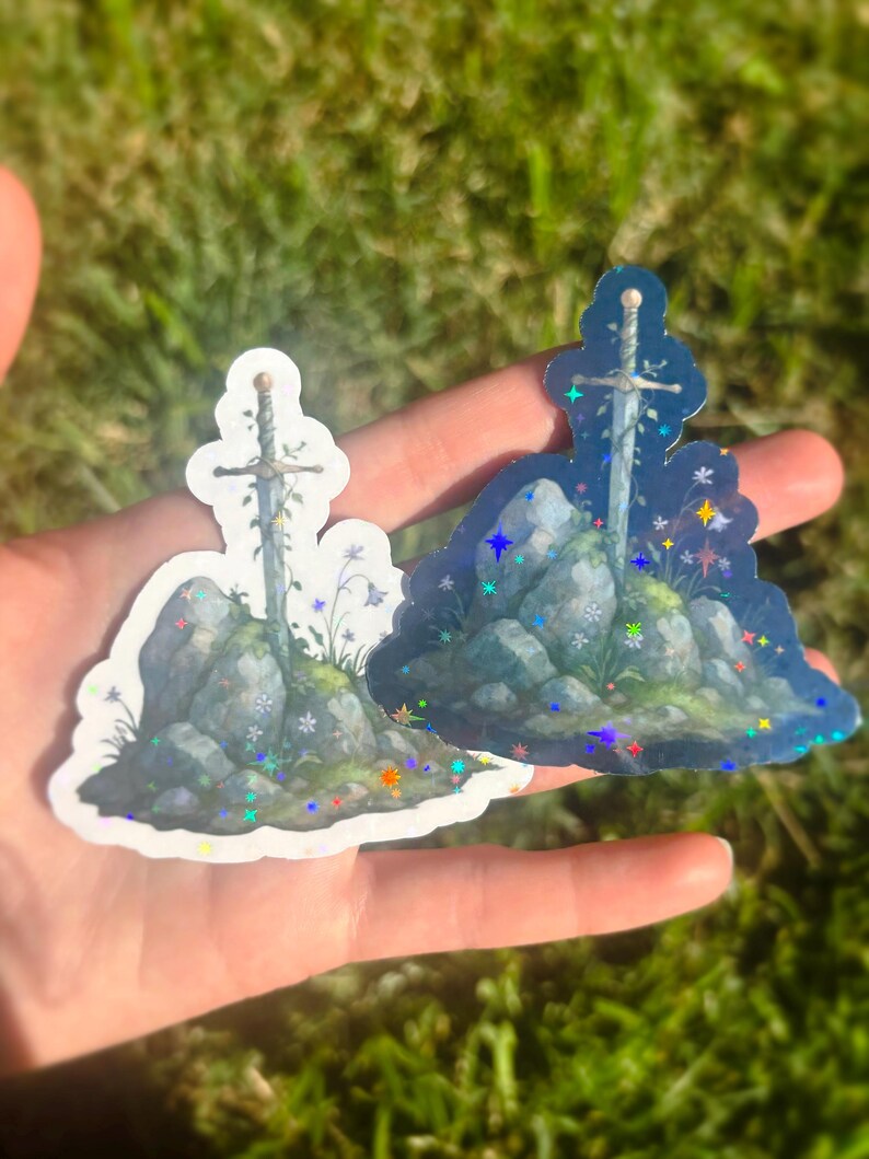 May include: Two stickers featuring a sword in a stone design. The stickers depict a sword embedded in a rock formation with floral accents. One sticker has a white background, and the other has a dark blue background with holographic glitter.