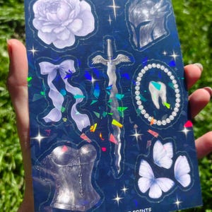 May include: A holographic sticker sheet featuring various fantasy-themed illustrations. Designs include a flower, bow, sword, helmet, tooth, corset, and butterflies. The text "PEACH CAT PRINTS" is visible at the bottom.