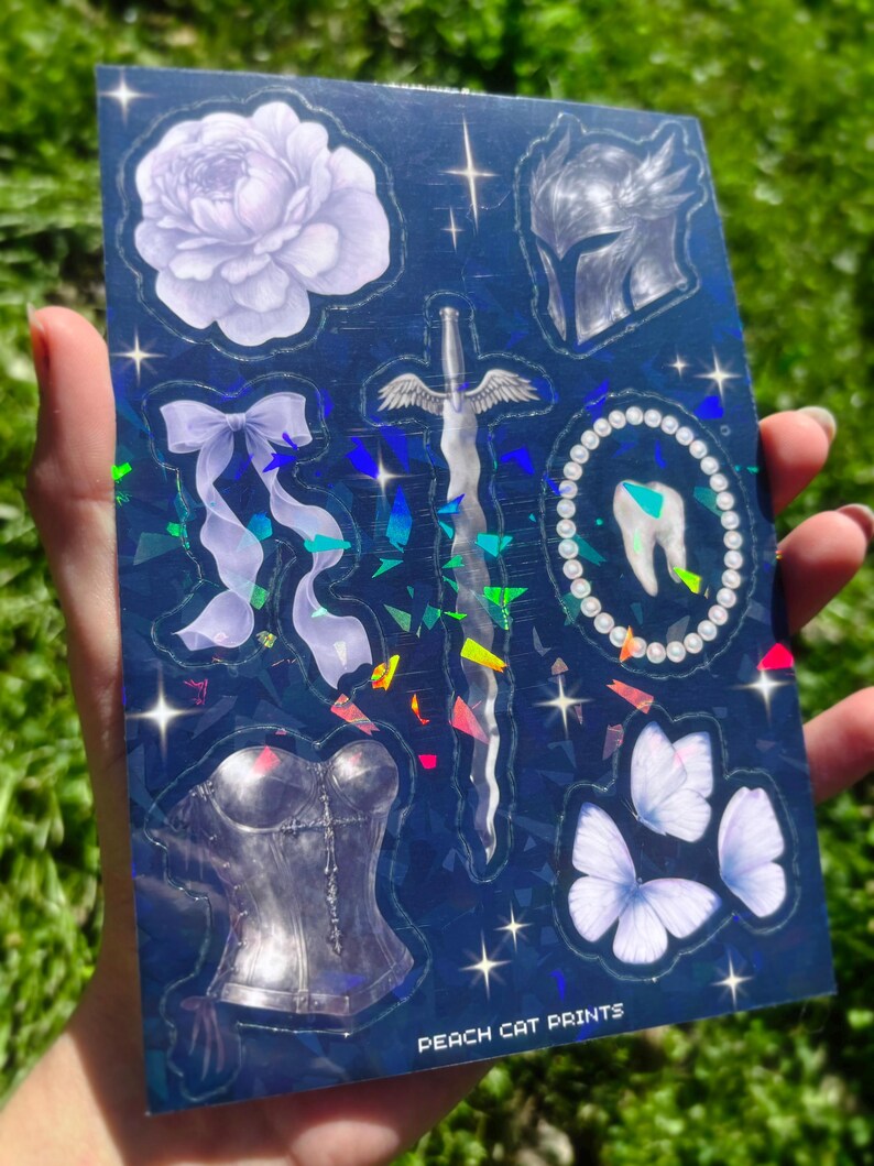May include: A holographic sticker sheet featuring various fantasy-themed illustrations. Designs include a flower, bow, sword, helmet, tooth, corset, and butterflies. The text "PEACH CAT PRINTS" is visible at the bottom.