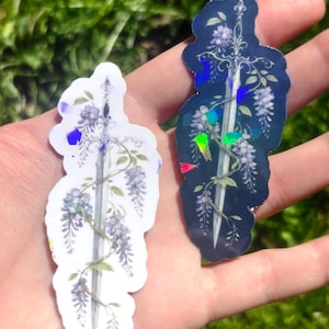May include: Two stickers featuring a sword design entwined with wisteria flowers. One sticker is white with purple accents, the other is navy blue with iridescent highlights. The stickers are held in a hand against a blurred green background.