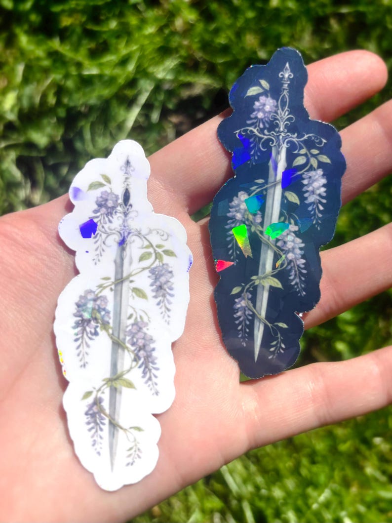 May include: Two stickers featuring a sword design entwined with wisteria flowers. One sticker is white with purple accents, the other is navy blue with iridescent highlights. The stickers are held in a hand against a blurred green background.