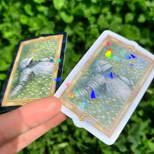Puede incluir: Dos pegatinas rectangulares con marco dorado y borde hologr&aacute;fico. Las pegatinas presentan una ilustraci&oacute;n de una armadura de caballero sobre un fondo floral verde. Las pegatinas se sostienen contra un fondo de follaje verde.