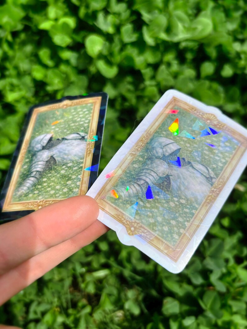 Puede incluir: Dos pegatinas rectangulares con marco dorado y borde hologr&aacute;fico. Las pegatinas presentan una ilustraci&oacute;n de una armadura de caballero sobre un fondo floral verde. Las pegatinas se sostienen contra un fondo de follaje verde.