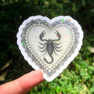 Pu&ograve; includere: Adesivo a forma di cuore con un'illustrazione di scorpione nero. L'adesivo ha uno sfondo bianco, un bordo di perle e un delicato bordo in pizzo. L'adesivo &egrave; tenuto su uno sfondo verde sfocato.