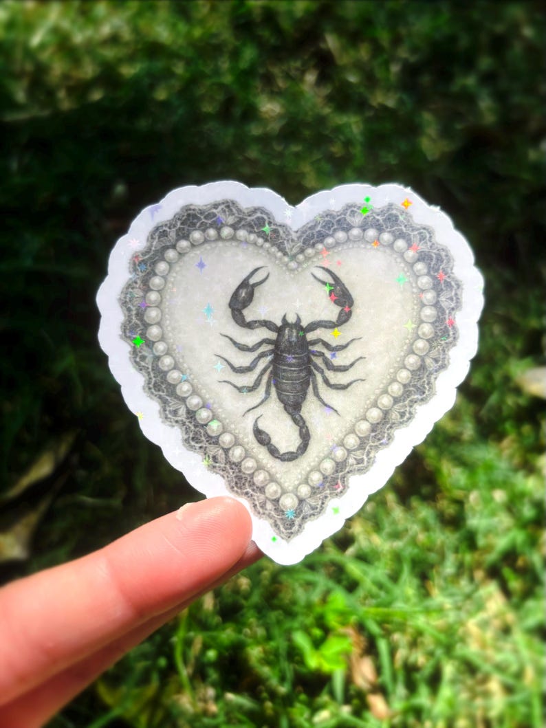 Pu&ograve; includere: Adesivo a forma di cuore con un'illustrazione di scorpione nero. L'adesivo ha uno sfondo bianco, un bordo di perle e un delicato bordo in pizzo. L'adesivo &egrave; tenuto su uno sfondo verde sfocato.
