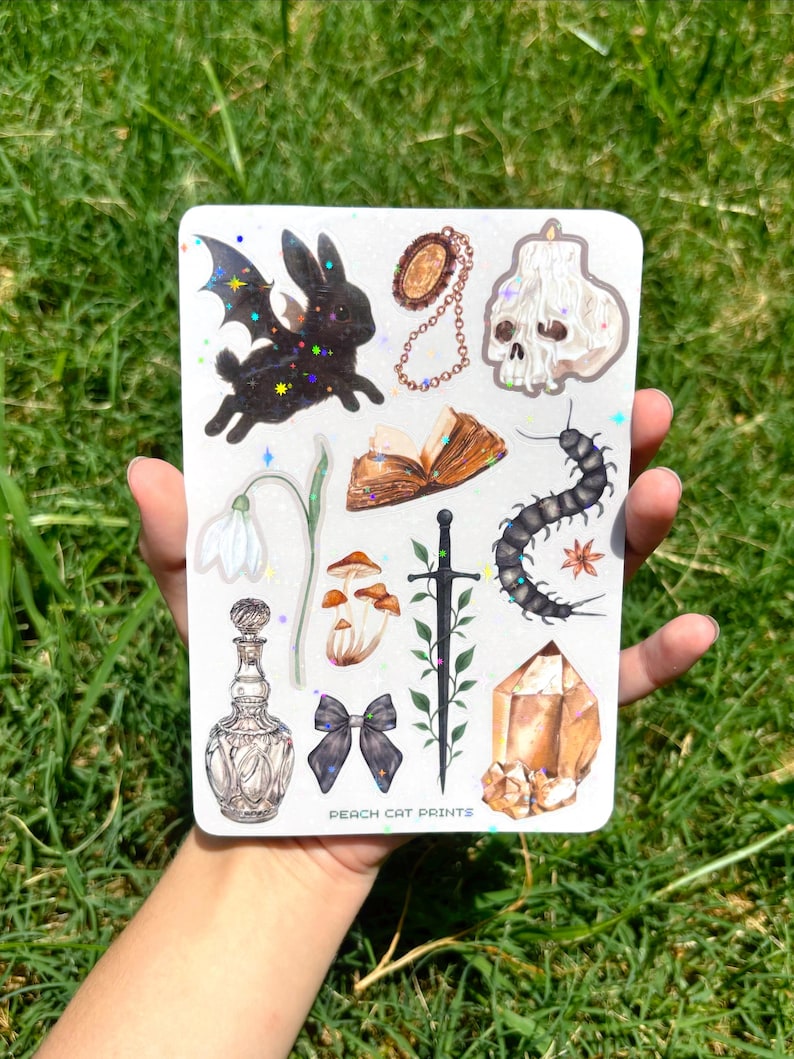 May include: A collection of stickers featuring various mystical and gothic-themed illustrations. Includes a black bat-winged rabbit, a skull candle, a crystal, a sword, a bow, and other designs. The text "PEACH CAT PRINTS" is visible.