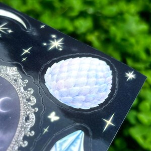 May include: A sheet of stickers with a celestial theme. The stickers feature a moon, stars, a crystal, and a round, feathery design in shades of blue and white. The background is a dark blue with a glossy finish.